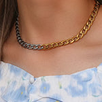 A close-up of a person's neck wearing a chunky chain necklace with alternating gold and silver-toned links, paired with a light blue and white floral top.