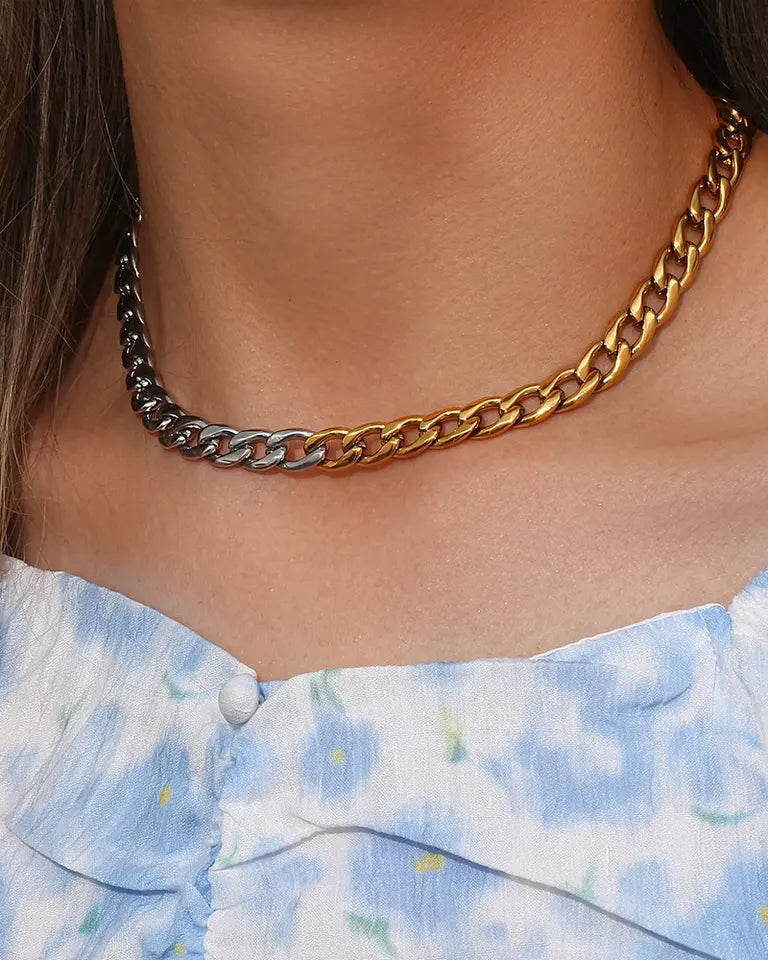 A close-up of a person's neck wearing a chunky chain necklace with alternating gold and silver-toned links, paired with a light blue and white floral top.