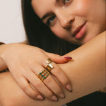 A close-up lifestyle shot of a woman wearing multiple gold rings, including one with green stones, on her hand resting on her arm.