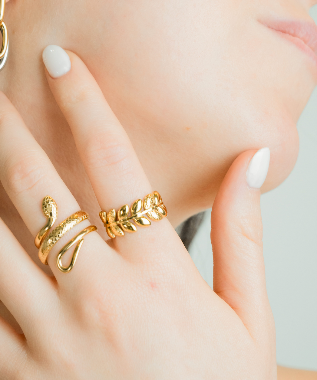 A close-up lifestyle shot of a model's hand and cheek, showcasing gold jewelry. The hand is adorned with a textured snake ring and a leaf-patterned ring, while the model's ear features a gold and silver hoop earring.