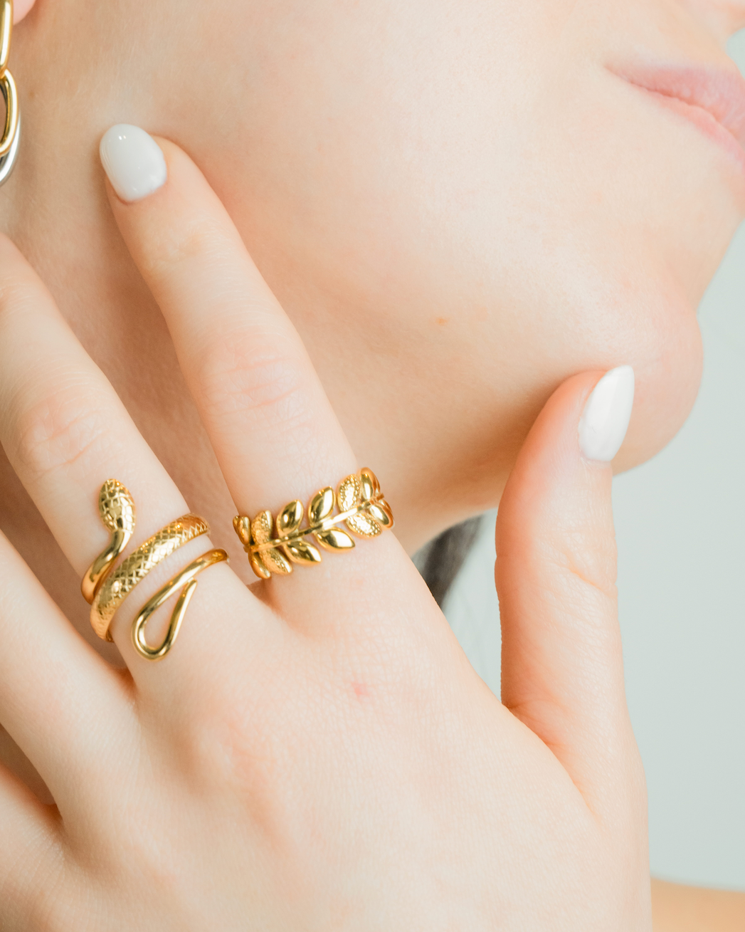 A close-up lifestyle shot of a model's hand and cheek, showcasing gold jewelry. The hand is adorned with a textured snake ring and a leaf-patterned ring, while the model's ear features a gold and silver hoop earring.