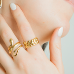 A close-up lifestyle shot of a model's hand and cheek, showcasing gold jewelry. The hand is adorned with a textured snake ring and a leaf-patterned ring, while the model's ear features a gold and silver hoop earring.