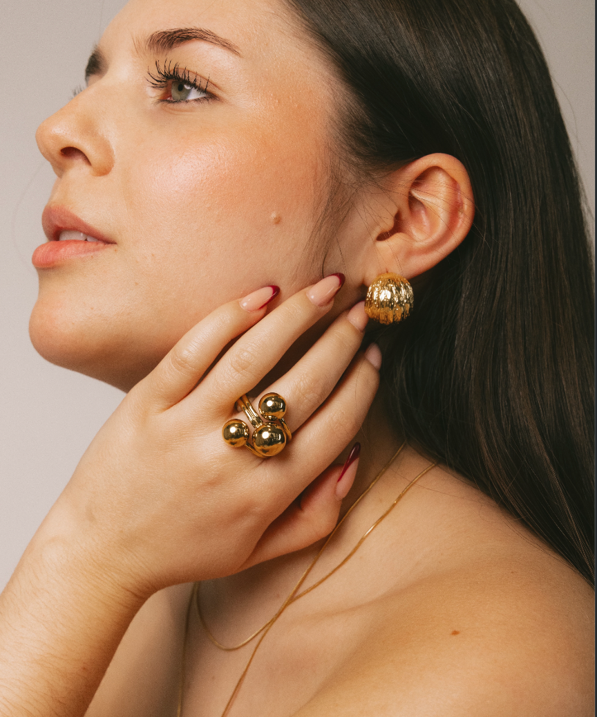 A close-up lifestyle shot of a woman wearing gold jewelry, featuring a chunky gold ring with three round beads on her finger and a matching gold textured earring on her ear.
