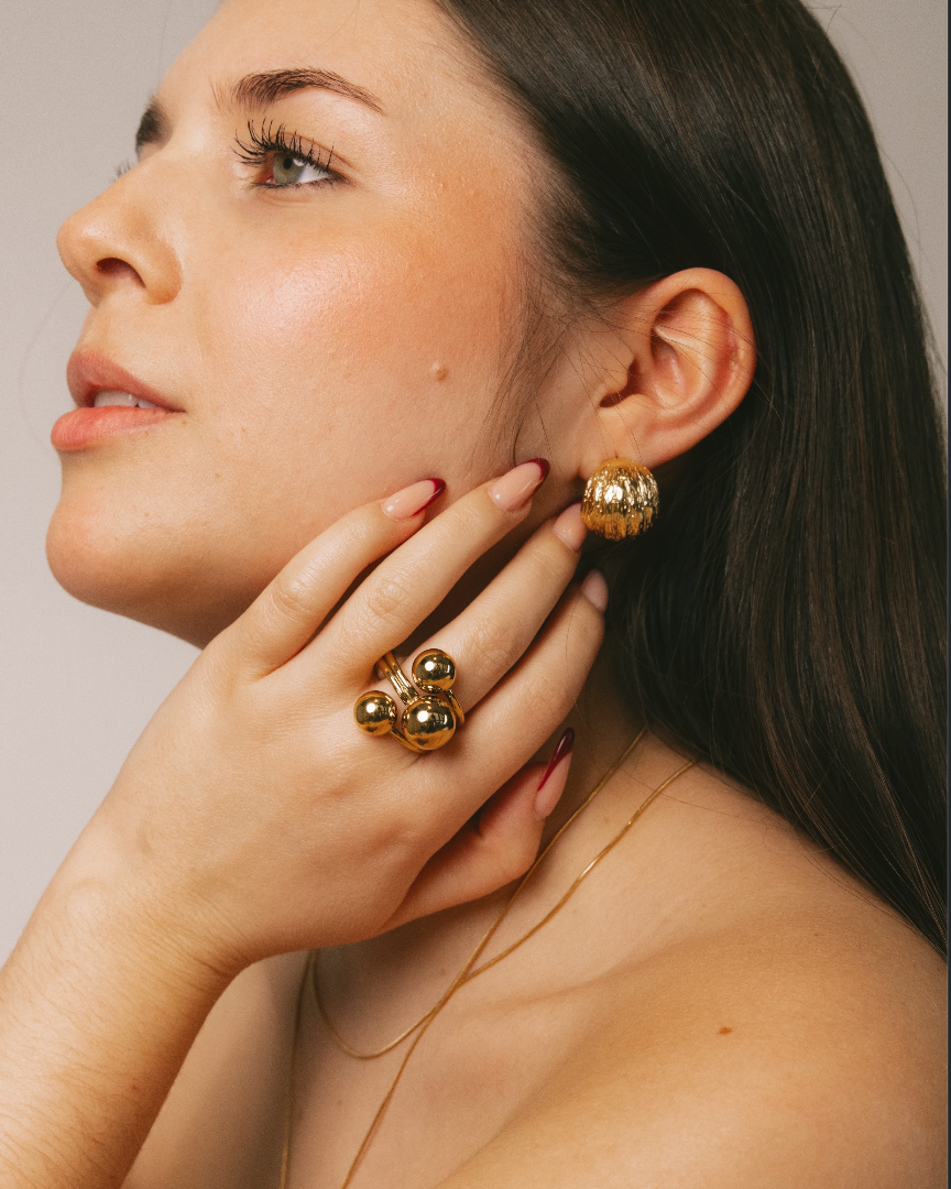 A close-up lifestyle shot of a woman wearing gold jewelry, featuring a chunky gold ring with three round beads on her finger and a matching gold textured earring on her ear.