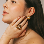 A close-up lifestyle shot of a woman wearing gold jewelry, featuring a chunky gold ring with three round beads on her finger and a matching gold textured earring on her ear.